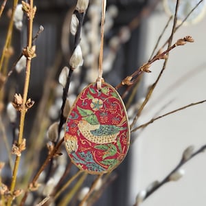 May include: An egg-shaped ornament with a red background and a bird design. The ornament is decorated with green leaves, strawberries, and a bird with blue and gold details. It hangs from a beige ribbon, perfect for Easter or spring decor.