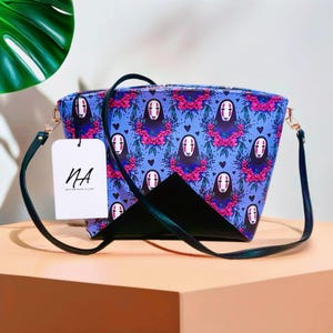 Puede incluir: Un bolso de hombro azul y negro con un patrón repetido de un personaje de cara blanca con flores rosas. El bolso tiene una correa negra y una etiqueta blanca con el texto "NA NOTORIOUS ALICE".