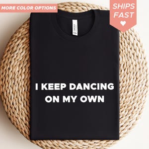 Puede incluir: Camiseta negra con el texto blanco "I KEEP DANCING ON MY OWN". La camiseta se muestra sobre una superficie tejida. Las palabras "MORE COLOR OPTIONS" y "SHIPS FAST" están en las esquinas superiores.
