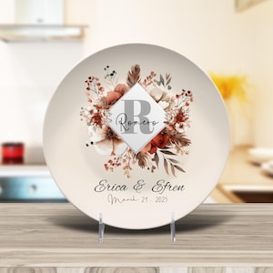 May include: A white ceramic plate with a floral design in shades of brown and red. The plate features a square with the letter "R" and the name "Romero" in black text. The text "Erica & Efrain March 21, 2025" is printed below the floral design.