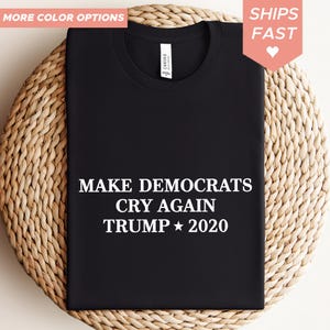 Puede incluir: Camiseta negra con texto blanco: "MAKE DEMOCRATS CRY AGAIN TRUMP 2020". La camiseta se muestra sobre una superficie tejida. La esquina superior izquierda tiene el texto "MÁS OPCIONES DE COLOR". La esquina superior derecha tiene un corazón rosa y las palabras "ENVÍOS RÁPIDOS".