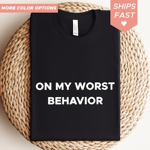Può includere: T-shirt nera con la scritta "ON MY WORST BEHAVIOR" a blocchi bianchi. La maglietta è esposta su una superficie tessuta. Le parole "MORE COLOR OPTIONS" e "SHIPS FAST" sono negli angoli superiori.