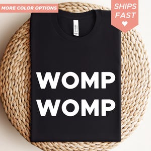 Può includere: T-shirt nera con la scritta bianca "WOMP WOMP". La maglietta è esposta su una superficie circolare intrecciata. Le parole "MORE COLOR OPTIONS" e "SHIPS FAST" sono negli angoli superiori.