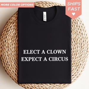 Può includere: T-shirt nera con la scritta bianca "ELECT A CLOWN EXPECT A CIRCUS". La maglietta è esposta su una superficie tessuta. Sono visibili anche le parole "MORE COLOR OPTIONS" e "SHIPS FAST".