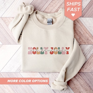 Könnte beinhalten: Ein beiger Pullover mit dem Text "HOLLY JOLLY web developer" in bunten, Retro-Lettern.