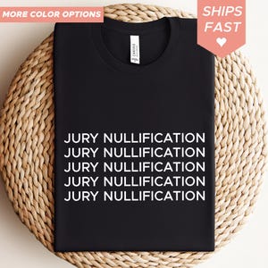 May include: Black t-shirt with the text "JURY NULLIFICATION" in white, repeated five times. The shirt is displayed on a woven, circular surface. The words "MORE COLOR OPTIONS" and "SHIPS FAST" are also visible.