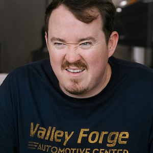 May include: A man wearing a navy blue t-shirt with the text "Valley Forge Automotive Center" in gold lettering. The man has a furrowed brow and a slight smile.