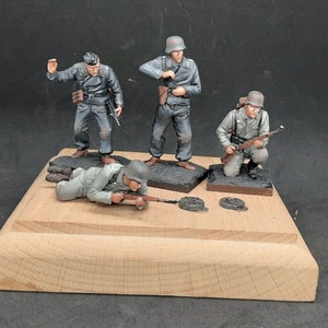 May include: Four painted miniature figures of soldiers in military uniforms, standing on a wooden base. The figures are holding rifles and are posed in a variety of action poses. The base is decorated with a small amount of dirt and grass.
