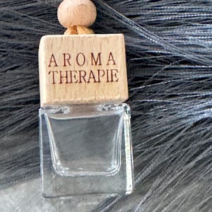 May include: A clear glass aromatherapy bottle with a wooden cap. The cap is engraved with the words "AROMA THERAPIE" in a dark brown font. The bottle is suspended by a tan cord and wooden bead.