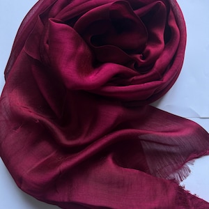 Può includere: Sciarpa bordeaux scuro con una texture morbida e fluida. Il tessuto è drappeggiato e arricciato, con un bordo sfrangiato visibile in basso. La sciarpa appare leggera e adatta alla stratificazione.