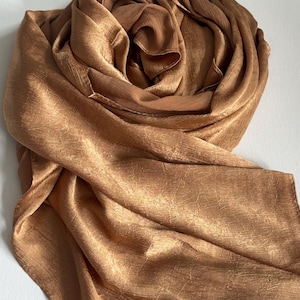 Puede incluir: Una bufanda de color bronce sólido con una superficie sutilmente arrugada y texturizada. La tela tiene un brillo metálico y está dispuesta en pliegues sueltos y fluidos, mostrando la calidad reflectante del material en toda su longitud.