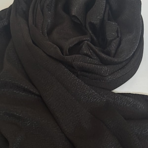 Puede incluir: Bufanda negra con superficie texturizada, drapeada y doblada. La tela tiene un brillo sutil, que capta la luz y crea reflejos y sombras. La bufanda parece suave y fluida, adecuada para varios estilos.