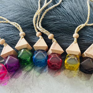 May include: A selection of colourful perfume diffusers with geometric shapes. Each diffuser has a wooden top and a faceted, jewel-toned glass container. The colours include pink, purple, green, blue, red, yellow, and brown. They hang from a beige cord.