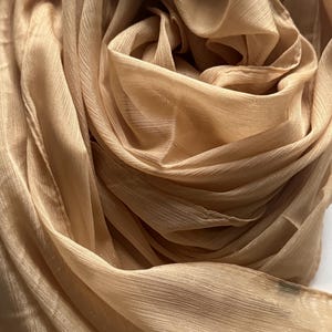 May include: Close-up of a beige scarf with a flowing, textured appearance. The fabric is gathered and draped, creating soft folds and shadows. The scarf appears to be made of a lightweight material, suitable for a variety of uses.