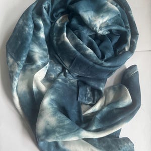 May include: A rolled, tie-dye scarf in shades of blue and white. The scarf is made of a soft, flowing fabric, with a pattern that resembles clouds or water. The scarf is a versatile accessory that can be worn in many ways.
