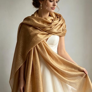 Könnte beinhalten: Ein schimmernder goldener Schal mit metallischem Glitzer-Finish, der über einem schlichten weißen ärmellosen Kleid liegt. Der leichte Stoff weist ein dezentes Funkeln auf und sorgt für ein weiches, fließendes Erscheinungsbild um die Schultern.