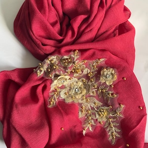 May include: A vibrant red scarf with an ornate gold floral design. The scarf is made of a soft fabric and features a detailed appliqué of gold beads and sequins. The design is centered on the scarf and adds a touch of elegance.
