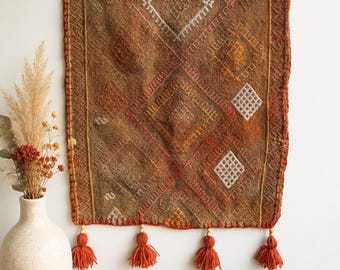 Könnte beinhalten: Ein quadratischer brauner Wandbehang aus Textil mit einem geometrischen Rautenmuster in Orange-, Beige- und Weißtönen. Die Kanten sind mit einem dicken orangefarbenen Schlingstich versehen, und am unteren Rand befinden sich vier orangefarbene Garnquasten mit Perlen.