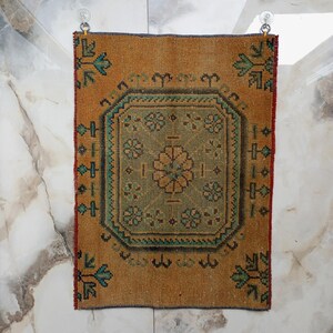 Pu&ograve; includere: Un piccolo tappeto rettangolare con un motivo geometrico centrale nei toni del verde e del beige, incorniciato da un bordo degli stessi colori. Il tappeto ha un aspetto invecchiato e vintage, con un bordo marrone rossiccio e accenti turchesi. &Egrave; appeso a due ventose.