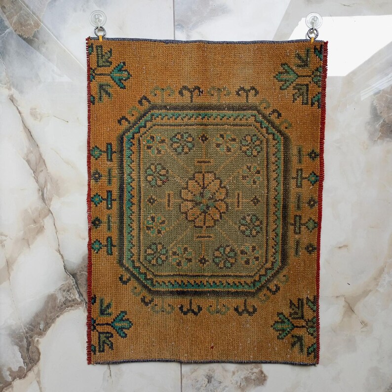 Pu&ograve; includere: Un piccolo tappeto rettangolare con un motivo geometrico centrale nei toni del verde e del beige, incorniciato da un bordo degli stessi colori. Il tappeto ha un aspetto invecchiato e vintage, con un bordo marrone rossiccio e accenti turchesi. &Egrave; appeso a due ventose.