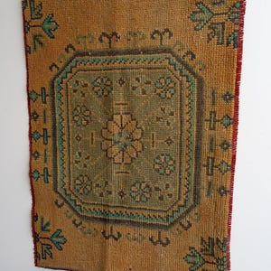Pu&ograve; includere: Un arazzo da parete rettangolare in stile vintage con un design geometrico centrale. Il tappeto presenta uno sfondo marrone dorato caldo con un motivo centrale verde e verde acqua. I bordi sono rifiniti con accenti rossi e blu, creando un aspetto rustico e strutturato.