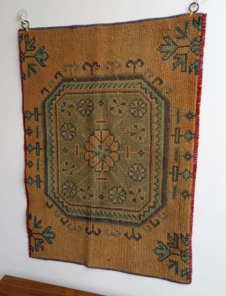 Pu&ograve; includere: Un arazzo da parete rettangolare in stile vintage con un design geometrico centrale. Il tappeto presenta uno sfondo marrone dorato caldo con un motivo centrale verde e verde acqua. I bordi sono rifiniti con accenti rossi e blu, creando un aspetto rustico e strutturato.