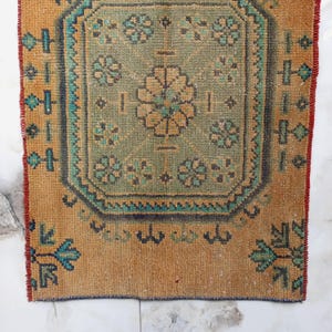 Pu&ograve; includere: Un piccolo tappeto rettangolare con un intricato motivo floreale sbiadito. Il tappeto presenta un disegno centrale nei toni del verde e dell'oro, incorniciato da un bordo turchese e blu scuro. I bordi sono di un caldo color marrone dorato con un bordo rosso.