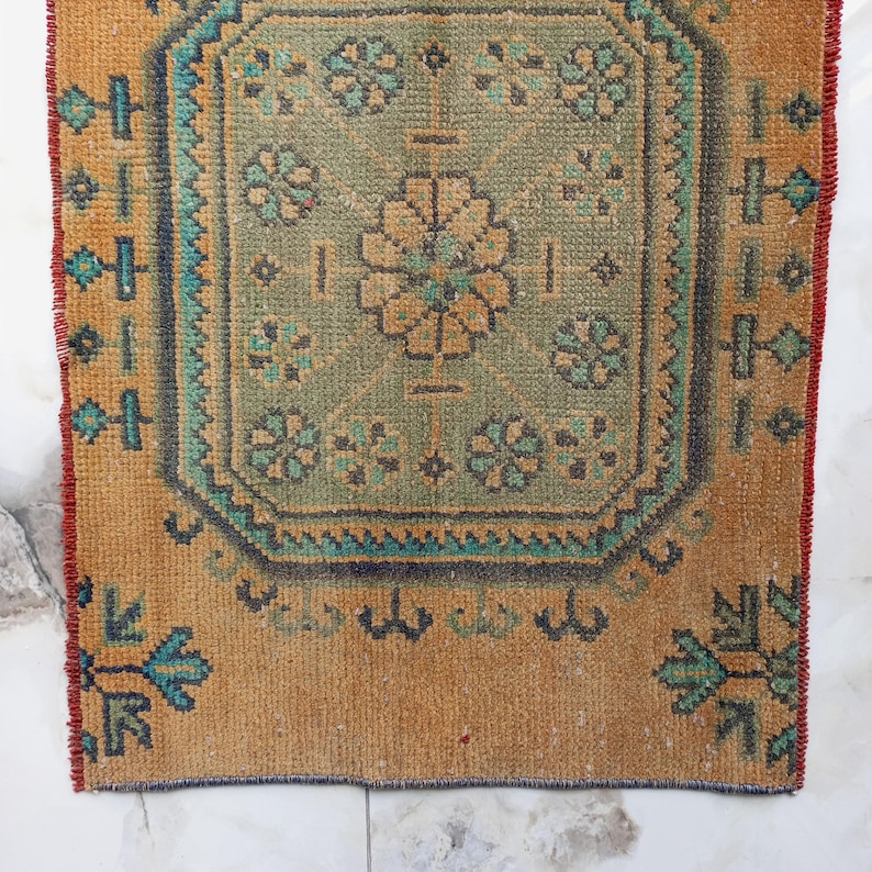Pu&ograve; includere: Un piccolo tappeto rettangolare con un intricato motivo floreale sbiadito. Il tappeto presenta un disegno centrale nei toni del verde e dell'oro, incorniciato da un bordo turchese e blu scuro. I bordi sono di un caldo color marrone dorato con un bordo rosso.