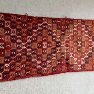 K&ouml;nnte beinhalten: Ein rot-wei&szlig;-schwarzer Teppich mit geometrischem Muster. Der Teppich ist mit komplizierten Details gewebt und h&auml;ngt an einer Wand.