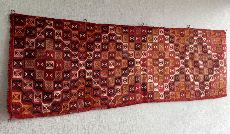 K&ouml;nnte beinhalten: Ein rot-wei&szlig;-schwarzer Teppich mit geometrischem Muster. Der Teppich ist mit komplizierten Details gewebt und h&auml;ngt an einer Wand.
