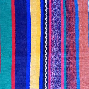 K&ouml;nnte beinhalten: Ein bunter gestreifter Teppich mit einer Vielzahl von Farben, darunter Gr&uuml;n, Rot, Blau, Gelb, Schwarz, Wei&szlig;, Rosa und Braun.