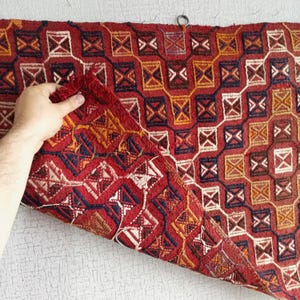 K&ouml;nnte beinhalten: Ein roter, blauer und gelber Teppich mit geometrischem Muster. Der Teppich ist in der Mitte gefaltet und wird von einer Hand gehalten.