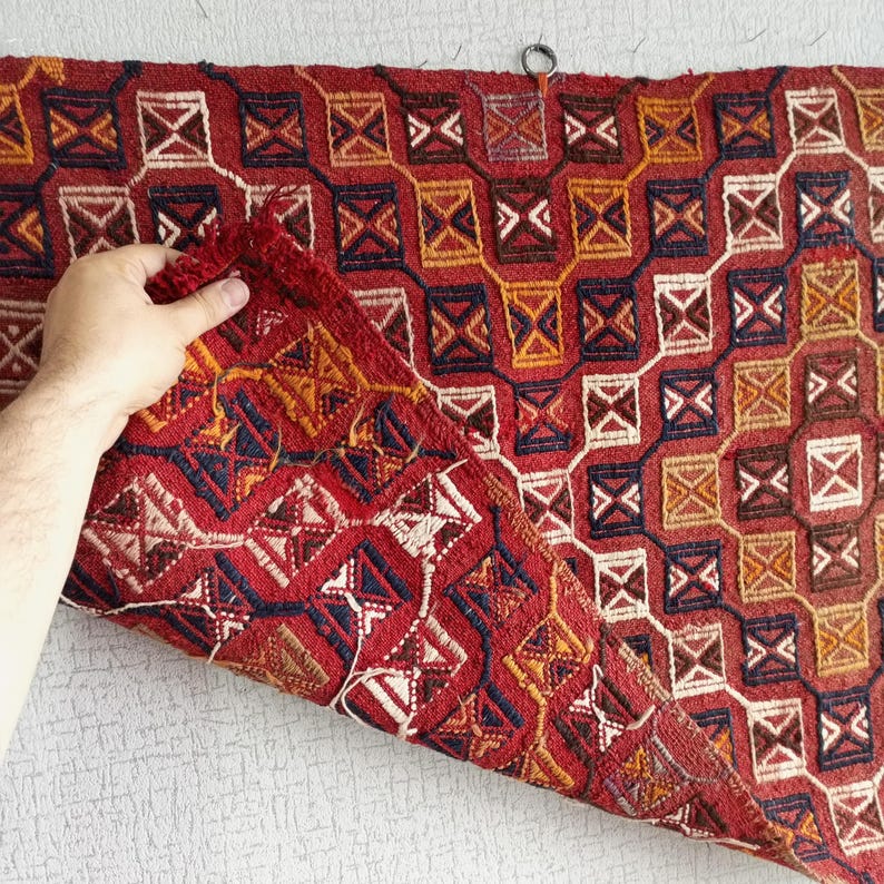 K&ouml;nnte beinhalten: Ein roter, blauer und gelber Teppich mit geometrischem Muster. Der Teppich ist in der Mitte gefaltet und wird von einer Hand gehalten.