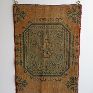 Pu&ograve; includere: Un tappeto rettangolare in stile vintage con un motivo floreale centrale verde e marrone. Il tappeto ha uno sfondo marrone con accenti turchesi e marroni. I bordi sono rifiniti con rosso e blu. Il tappeto &egrave; appeso a una parete bianca.