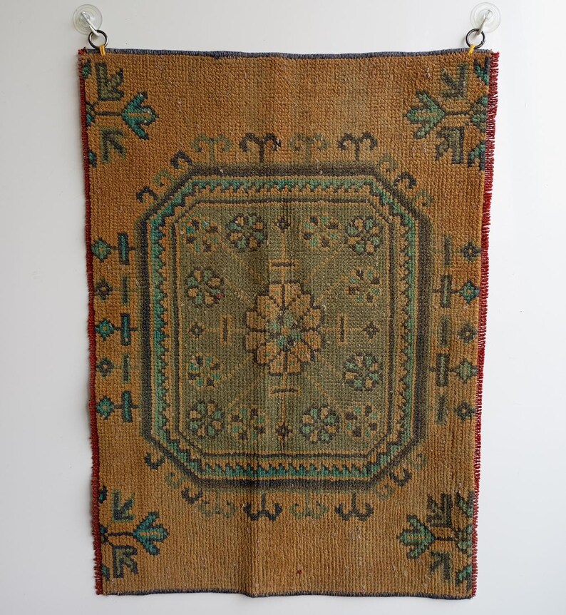 Pu&ograve; includere: Un tappeto rettangolare in stile vintage con un motivo floreale centrale verde e marrone. Il tappeto ha uno sfondo marrone con accenti turchesi e marroni. I bordi sono rifiniti con rosso e blu. Il tappeto &egrave; appeso a una parete bianca.