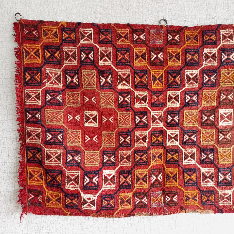 K&ouml;nnte beinhalten: Ein roter, orangefarbener und blauer geometrisch gemusterter Teppich mit Fransen an allen Seiten. Der Teppich h&auml;ngt an einer wei&szlig;en Wand.