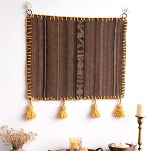 May include: A rectangular brown wall hanging with vertical stripes and a yellow stitched border. The wall hanging has four yellow tassels at the bottom. It is mounted on a white wall with two suction cups.