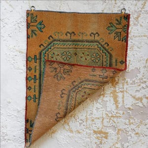 Pu&ograve; includere: Un piccolo tappeto rettangolare vintage nei toni dell'arancione e del marrone. Presenta intricati motivi geometrici in verde acqua e verde, con un bordo rosso. Due anelli di metallo sono attaccati per appenderlo.
