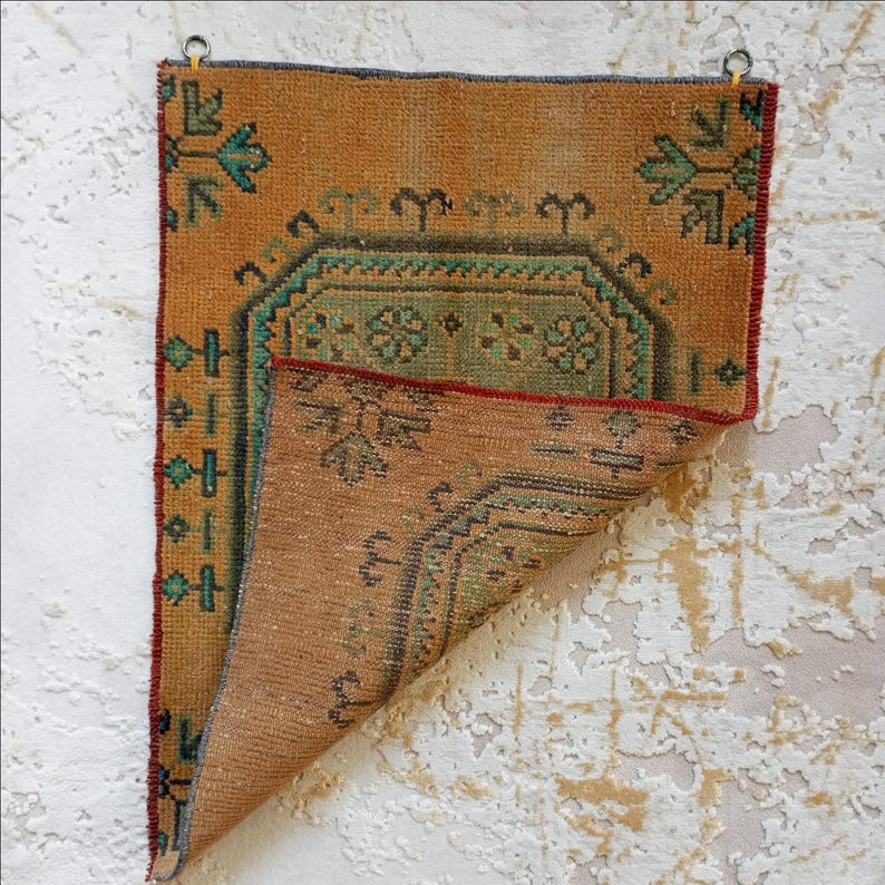 Pu&ograve; includere: Un piccolo tappeto rettangolare vintage nei toni dell'arancione e del marrone. Presenta intricati motivi geometrici in verde acqua e verde, con un bordo rosso. Due anelli di metallo sono attaccati per appenderlo.