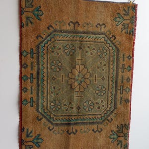 Pu&ograve; includere: Un tappeto rettangolare in stile vintage con un motivo geometrico centrale nei toni del verde e del marrone. Il tappeto ha uno sfondo marrone con accenti turchese e blu scuro. I bordi sono rifiniti con rosso e blu scuro. Il tappeto &egrave; appeso a una parete bianca.