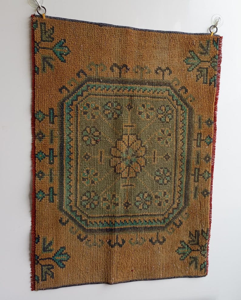 Pu&ograve; includere: Un tappeto rettangolare in stile vintage con un motivo geometrico centrale nei toni del verde e del marrone. Il tappeto ha uno sfondo marrone con accenti turchese e blu scuro. I bordi sono rifiniti con rosso e blu scuro. Il tappeto &egrave; appeso a una parete bianca.
