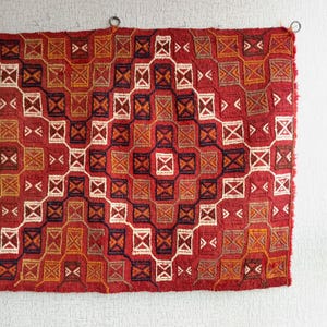 K&ouml;nnte beinhalten: Ein roter, orangefarbener und wei&szlig;er gewebter Teppich mit geometrischem Muster. Der Teppich hat einen Fransenrand und h&auml;ngt an einer wei&szlig;en Wand.