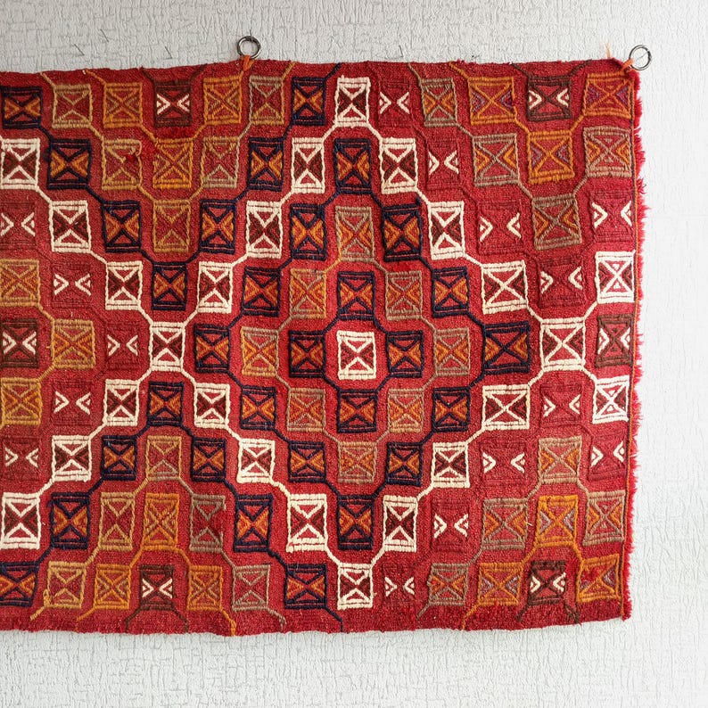 K&ouml;nnte beinhalten: Ein roter, orangefarbener und wei&szlig;er gewebter Teppich mit geometrischem Muster. Der Teppich hat einen Fransenrand und h&auml;ngt an einer wei&szlig;en Wand.