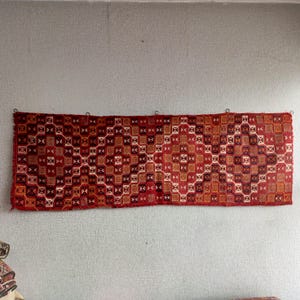 K&ouml;nnte beinhalten: Ein rot-wei&szlig;-brauner Teppich mit geometrischem Muster. Der Teppich h&auml;ngt an einer Wand.