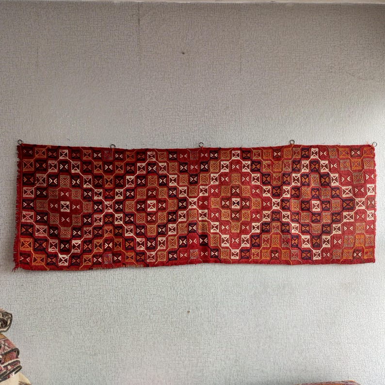 K&ouml;nnte beinhalten: Ein rot-wei&szlig;-brauner Teppich mit geometrischem Muster. Der Teppich h&auml;ngt an einer Wand.