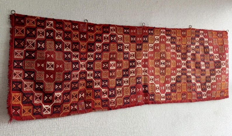 K&ouml;nnte beinhalten: Ein rot-wei&szlig;-schwarzer Teppich mit geometrischem Muster. Der Teppich ist mit komplizierten Details gewebt und h&auml;ngt an einer Wand.