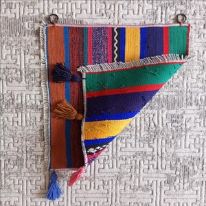 K&ouml;nnte beinhalten: Ein bunter gewebter Wandbehang mit Fransen und Quasten. Der Behang zeigt eine Vielzahl von Farben, darunter Rot, Blau, Gelb, Gr&uuml;n und Lila. Der Behang ist in der Mitte gefaltet, sodass die R&uuml;ckseite sichtbar ist.