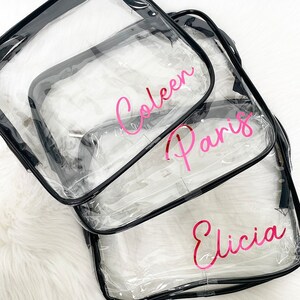 May include: Three clear plastic zippered bags with black trim. The bags are stacked on top of each other. The top bag has the name "Coleen" written in pink lettering. The middle bag has the name "Paris" written in pink lettering. The bottom bag has the name "Eliccia" written in pink lettering.