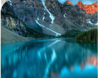 May include: A scenic landscape featuring a turquoise lake reflecting the surrounding mountains. The mountain peaks are illuminated with golden sunlight, contrasting with the dark, rocky slopes and patches of snow. A forest lines the lake's edge.