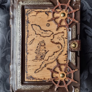 Puede incluir: Un diario encuadernado en cuero marrón con una cubierta de madera que presenta un mapa y un diseño de barco. El diario tiene dos ruedas de barco de madera decorativas en la portada.